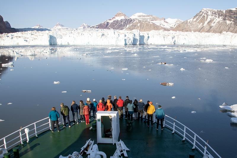 Folk står i baugen på ein båt. Dei ser på ein isbre som ligg ned mot havet, fjell i bakgrunnen.